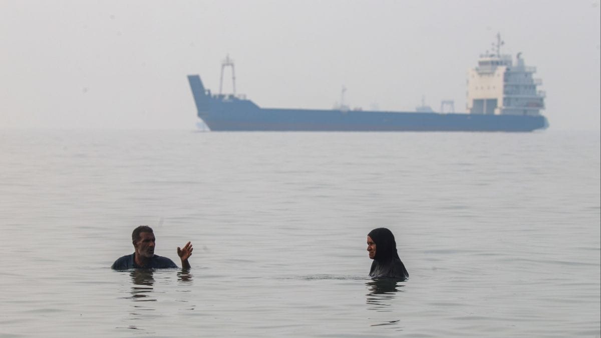 A man and a woman stand in shallow water as a commercial vessel appears anchored in the Strait of Hormuz off Bandar Abbas, Iran on Oct, 6, 2026 | AP