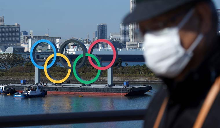 A man wearing a protective face mask to help curb the spread of the coronavirus walks with the Olympic rings in the background in the Odaiba section, in Tokyo | AP