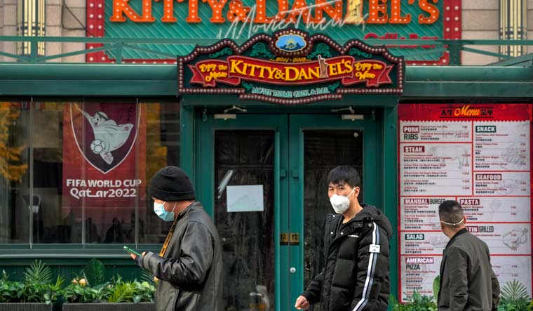 People wearing face masks walk by a shuttered restaurant displaying posters promoting the Qatar FIFA World Cup, as part of COVID-19 controls in Beijing, Tuesday, Nov. 22, 2022 | AP
