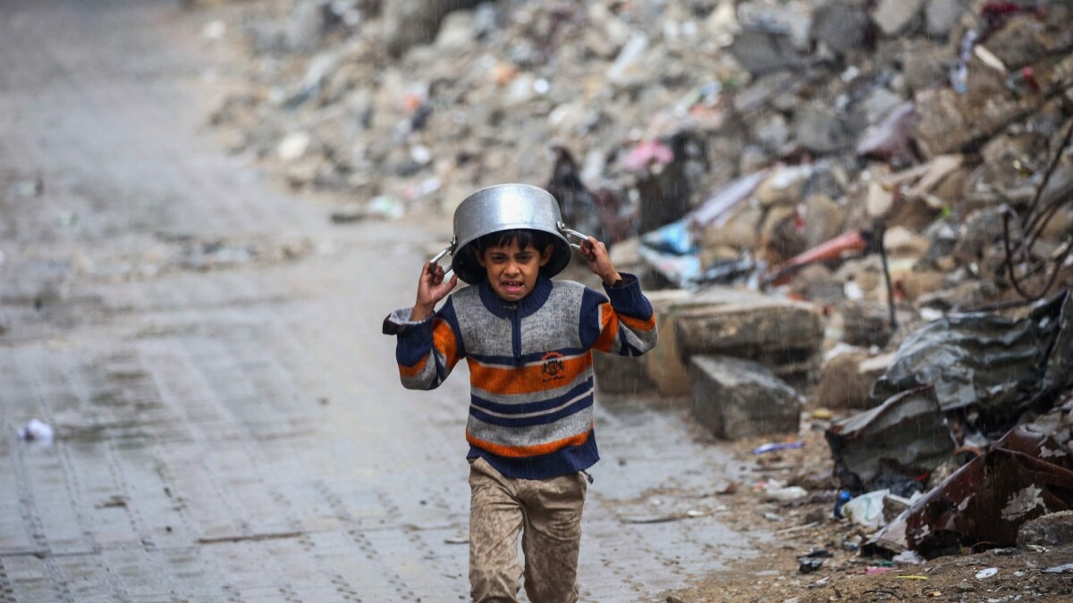 A displaced boy covers his head with a pan as he runs from the rain past building rubble at the Bureij refugee camp in the central Gaza Strip | AFP