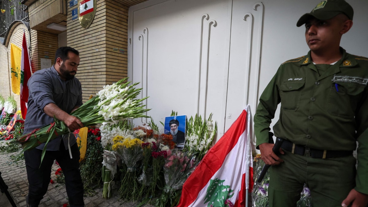 A member of the Iranian security forces stands guard as a man places flowers at the site of a memorial set up outside Lebanon's embassy in Tehran | AFP
