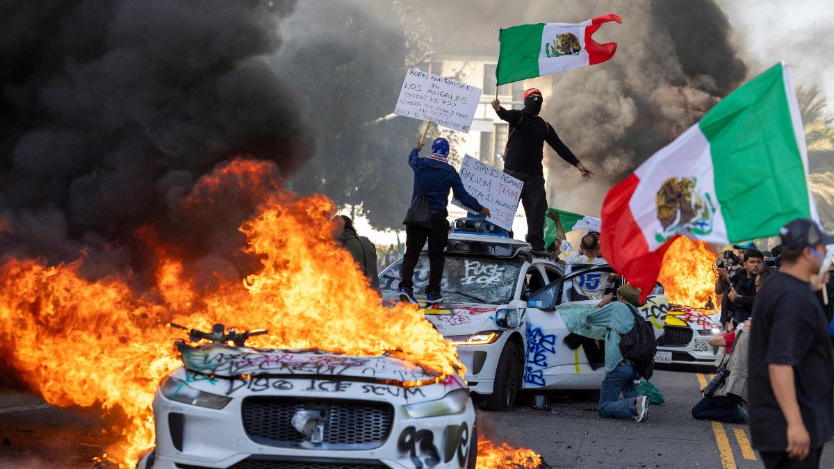 Demonstrators wave flags from atop a wrecked car, standing beside burning Waymo vehicles, during a protest in Los Angeles, California, on June 8, 2025 | AFP
