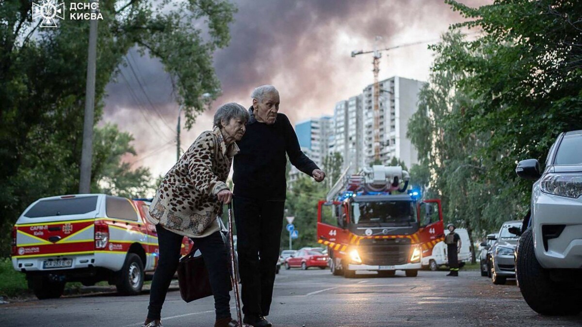 An elderly couple crossing a street as smoke rises in the background at a site of a strike following a mass Russian drone and missile attack on Kyiv | AFP